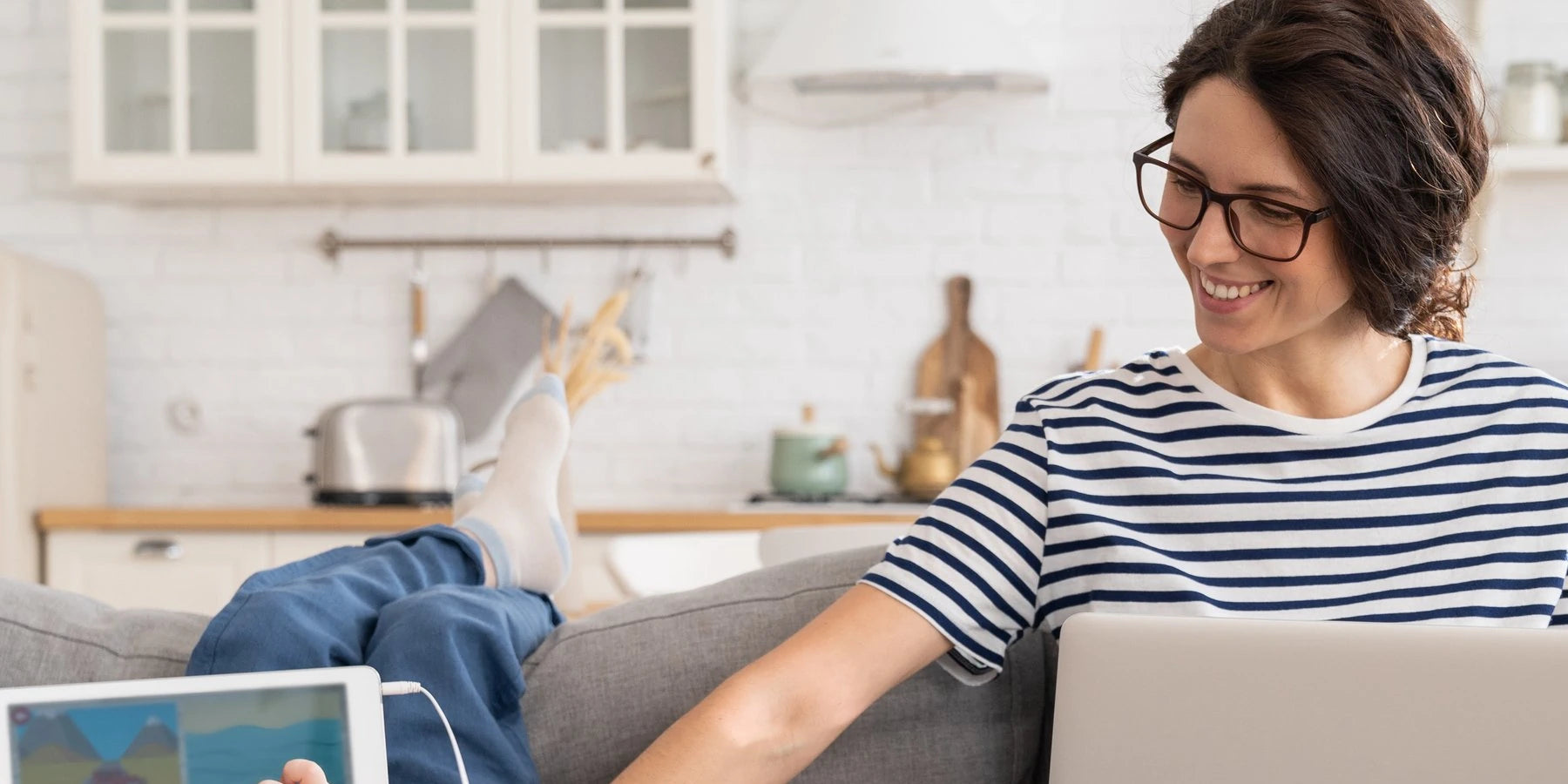 Person wearing glasses while sitting on a couch, legs crossed with a laptop.